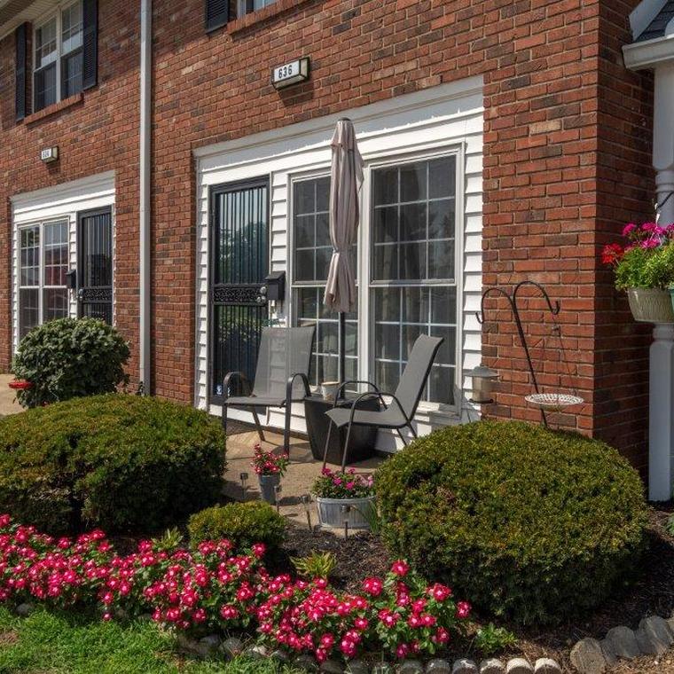 Apartment patio with beige umbrella and patio furniture surrounded by trimmed bushes and pink flowers